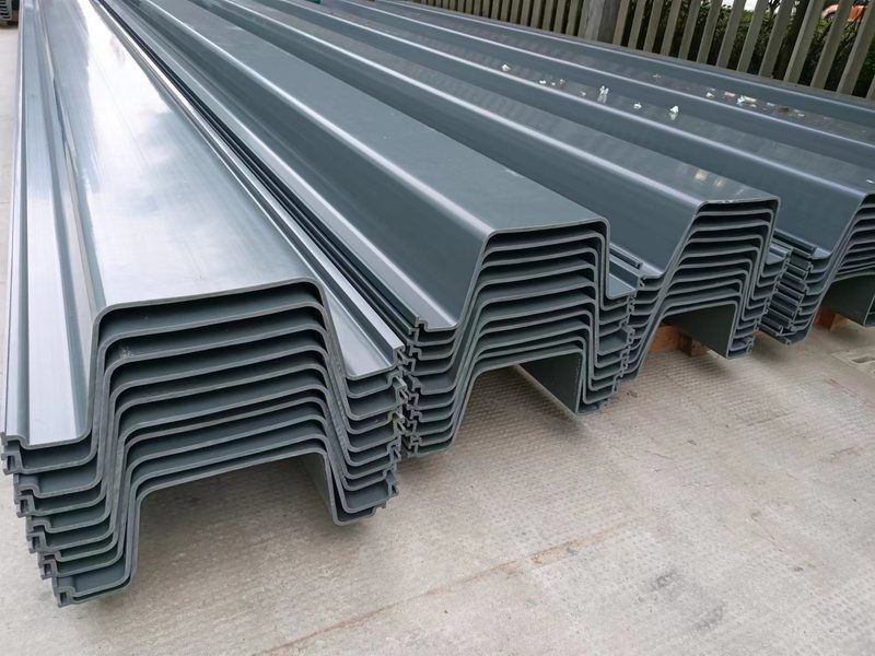 Gray PVC sheet pile profiles stacked on timber spacers with profile ends and interlocks visible. Gray PVC sheet pile profiles stacked on timber spacers with profile ends and interlocks visible.